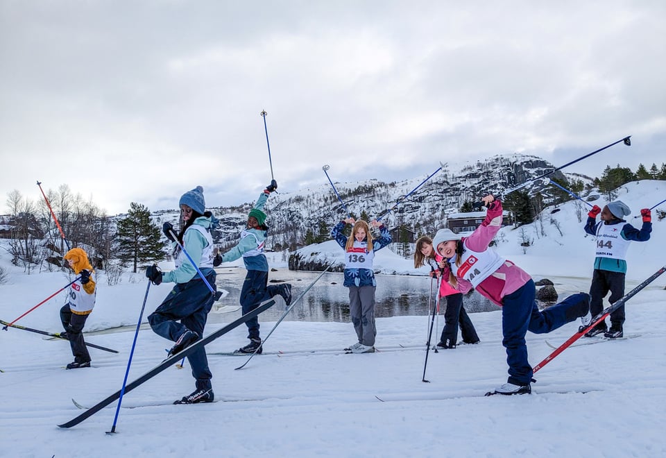 En gjeng med barn som står på langrennski vifter opp med skiene og stavene og smiler til kamera.