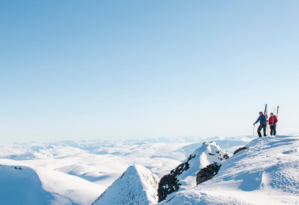 To personer med ski på sekken på en snødekket fjelltopp