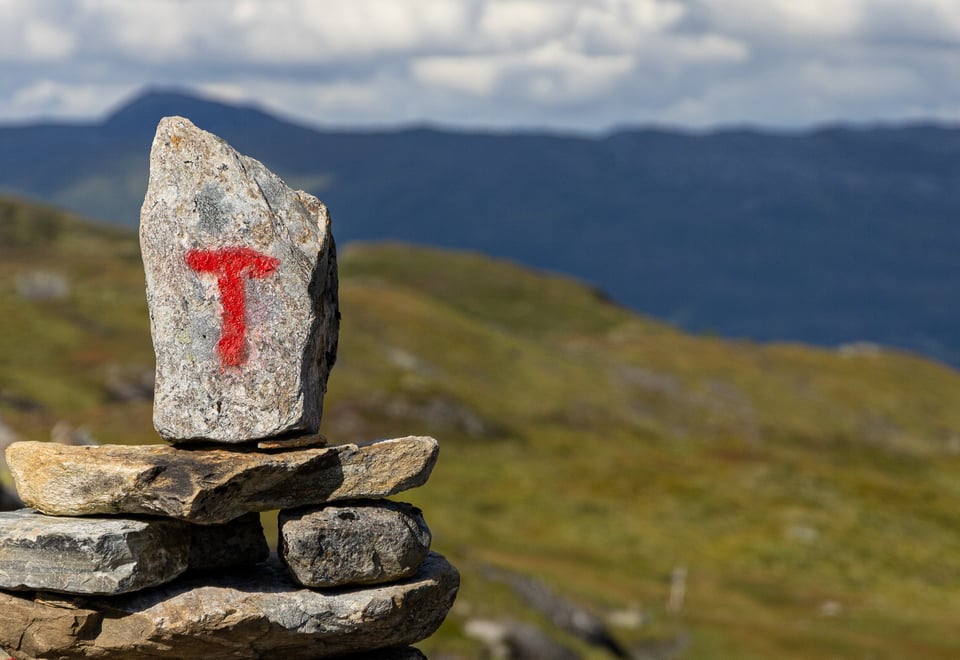 Rødmalt T på varde med fjell og lette skyer i bakgrunnen.