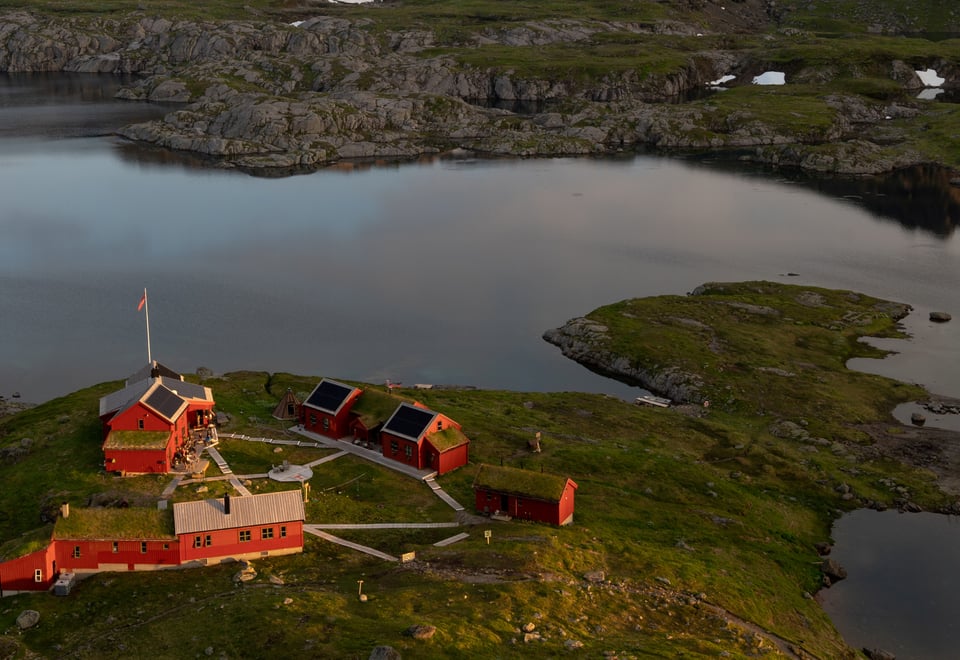 Dronebilde av hytta med oversikt over hytta, vannet og fjellene.