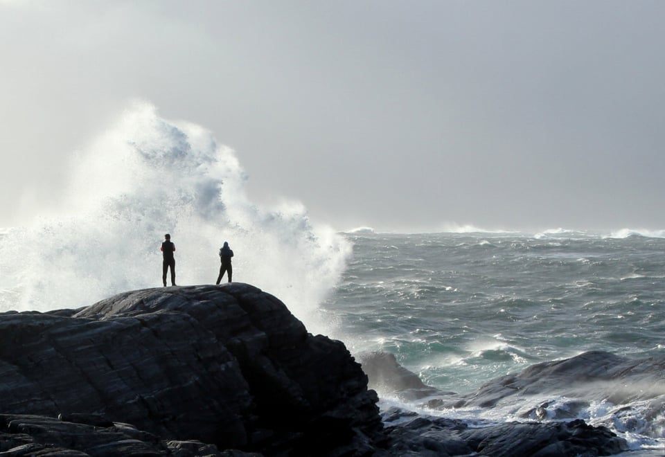 To mennesket står på svaberg og ser på store bølger ute i havgapet. Foto