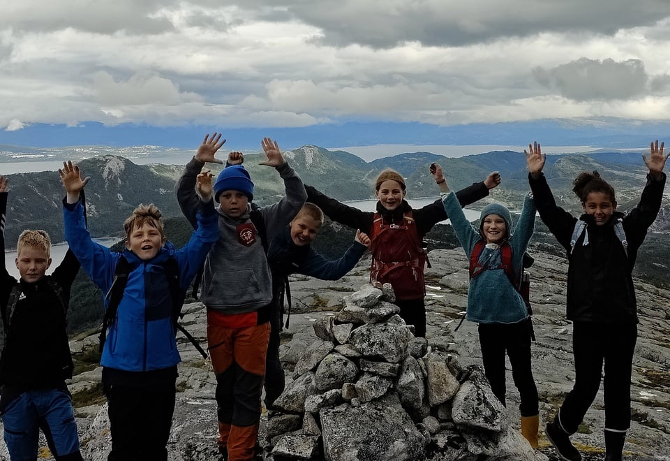 En gjeng ungdommer på toppen av fjellet med hendene opp i været og varden foran seg.