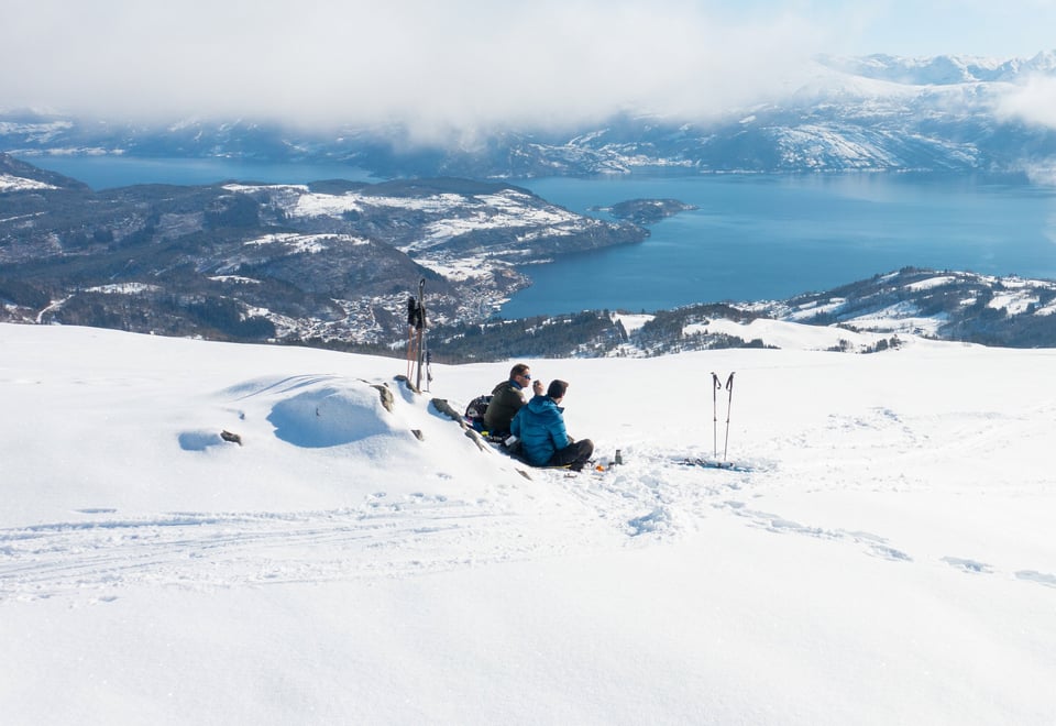 To personer har pause i en bakke med snø. Sol og fin utsikt over Hardangerfjorden.