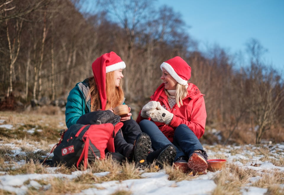 To damer sitter i snødekt gress med nisseluer. De ler og koser seg mens de drikker kaffe og spiser pepperkaker.