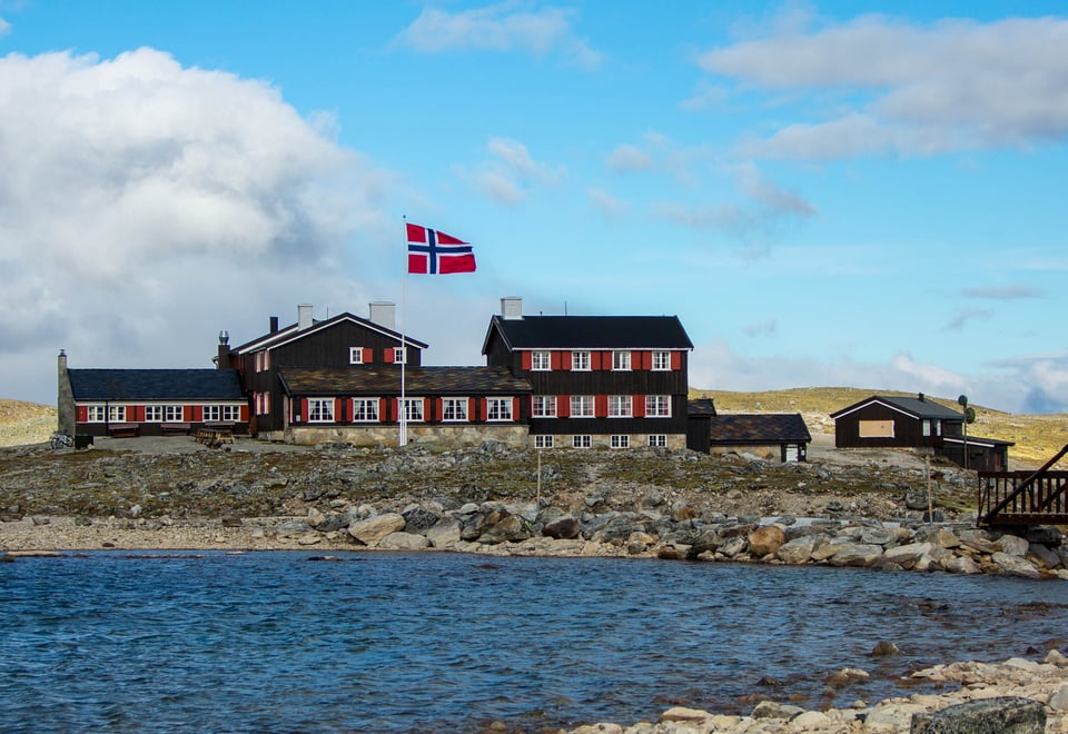 Svartmalt hytte med rød karmer og en flaggstang med norsk flagg foran hytta