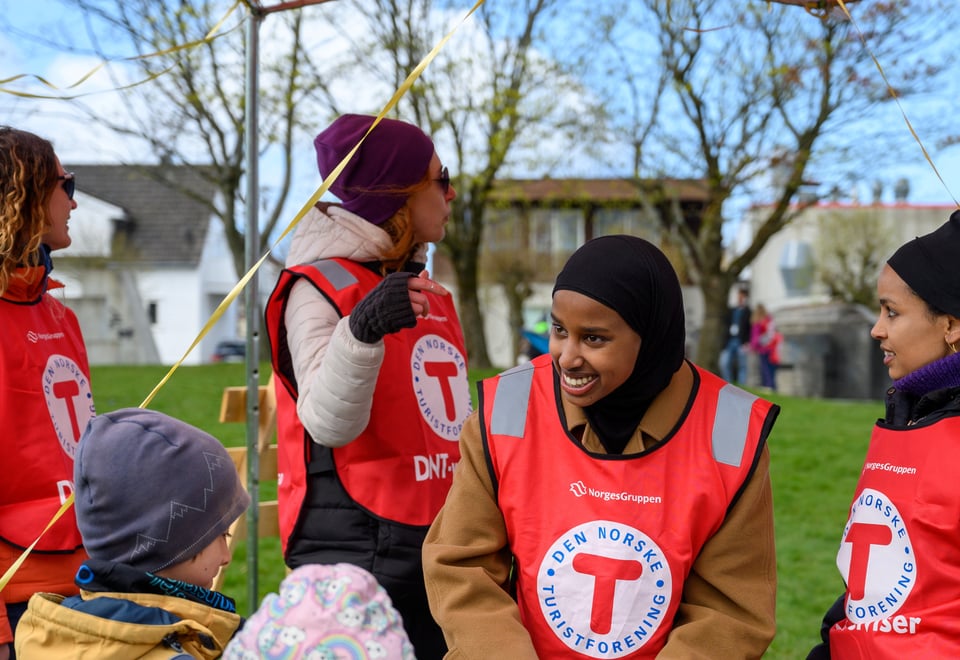 En dame med sjal og rød DNT vest smiler til et barn som står med ryggen til. Noen andre damer med rød DNT vest står rundt henne. 