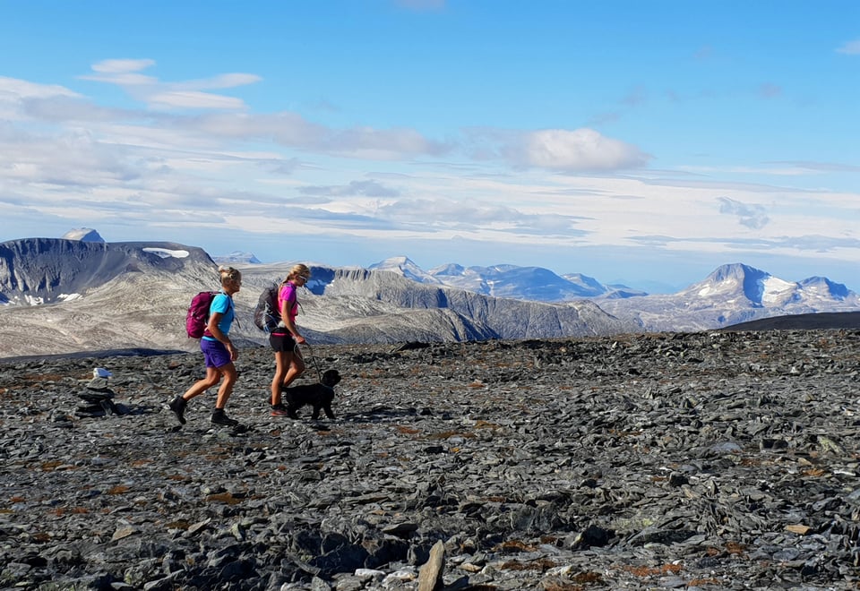 To kvinner og en liten hund i bånd som går over en steinete og øde landskap, med store fjell i bakgrunnen