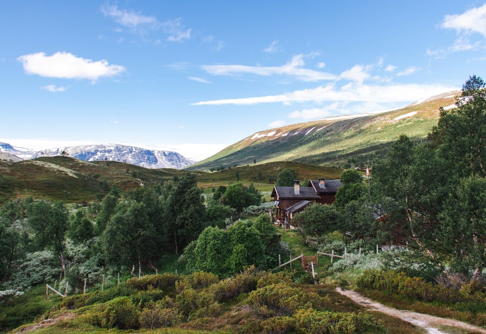 Bildet viser en åpen fjelldal med mye lyngterreng og noe buskvekst. En sti fører ned til en liten hytte, og i bakgrunnen ruver store fjell.