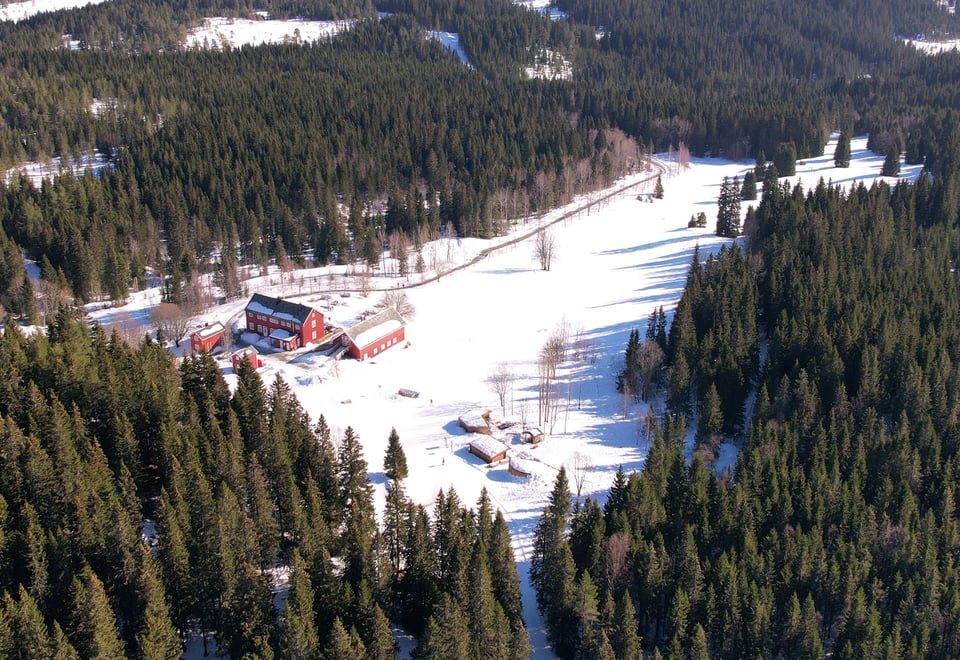 Dronebilde av en klynge rødmalte bygninger sett langt ovenfra på vinterstid, med skoglandskapet rundt