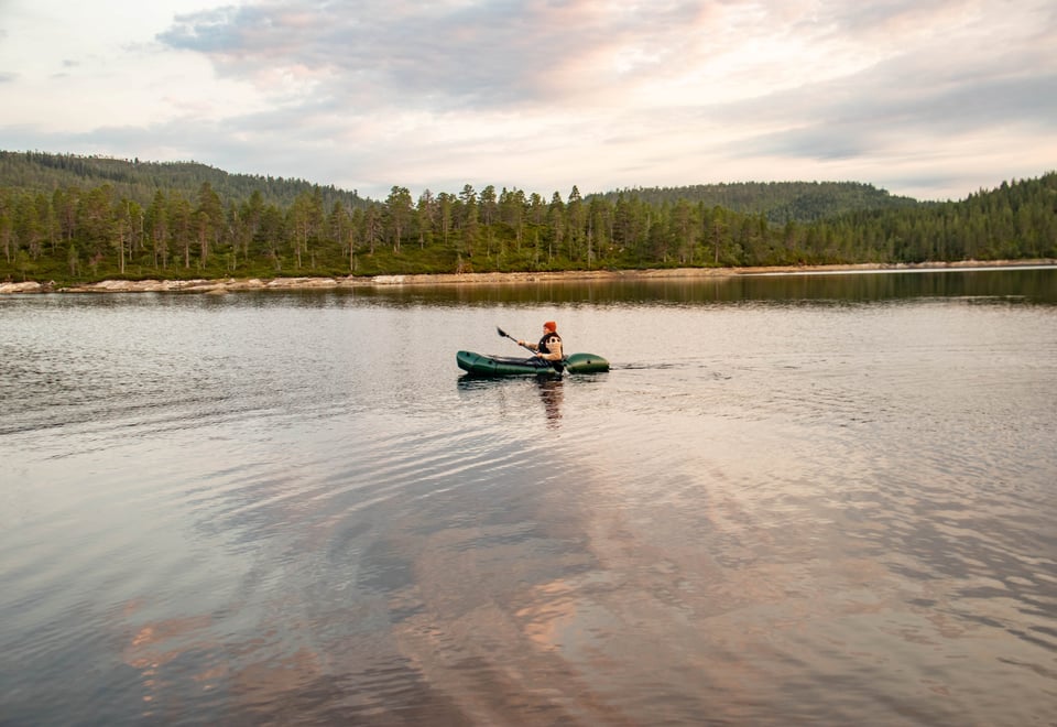 Kvinne padler alene i en packraft på en innsjø, med trær langs vannbredden