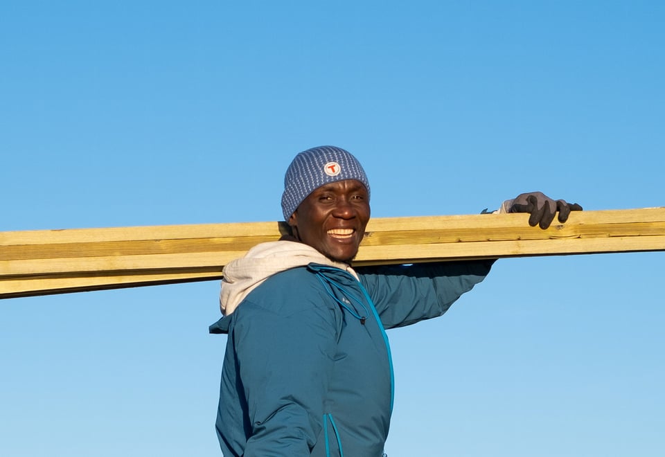 Smilende mann med grønn jakke og blå lue  holder noen planker mens det er blå himmel i bakgrunnen