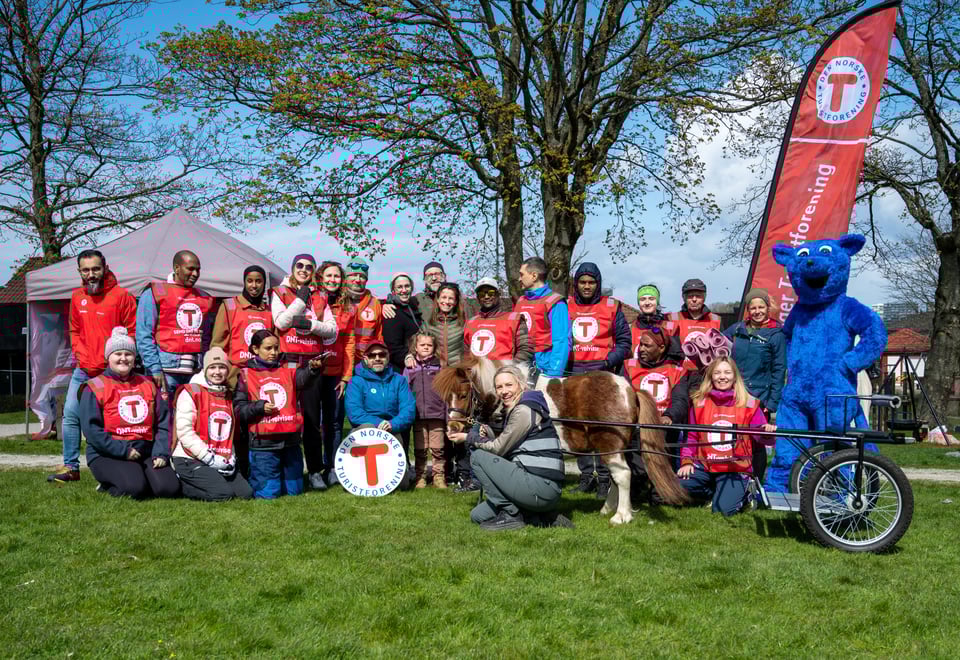 Mange smilende mennesker med røde DNT vester står sammen med en stor blå bamse og DNT beach flagg.