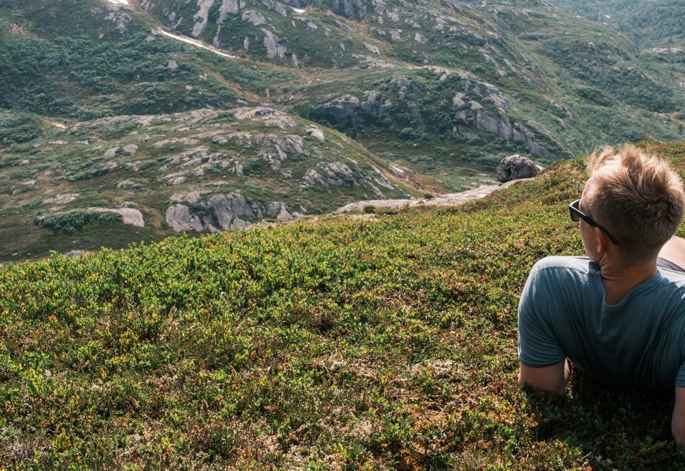 En mann som har solbriller, shorts, t-skjorte og fjellsko, ligger i gresset og ser utover fjellene