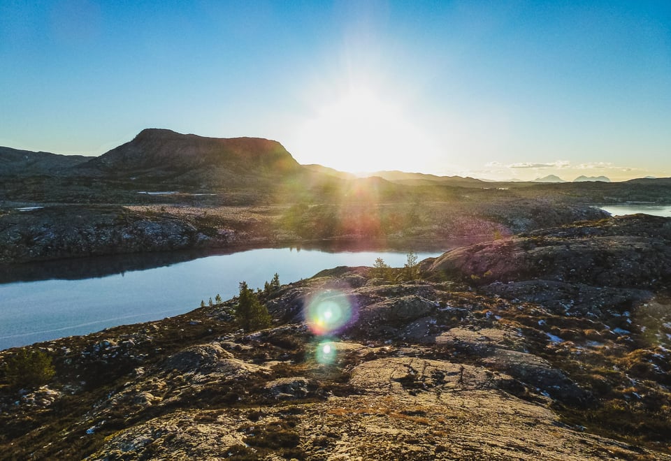 Åpent og steinete landskap delt av vann mellom forgrunn og bakgrunn. Et lavt og bredt fjell står foran horisonten midt i bildet, med solen rett til høyre for seg. Noen få spredte små planter her og der. En rekke fjell strekker seg ut lenger og lenger unna i det fjerne.