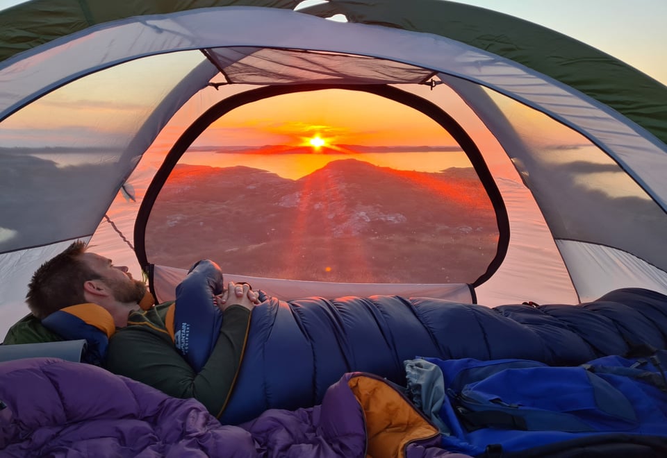 Bilde av innsiden av et telt, hvor en mann ligger i sovepose og ser ut gjennom bakveggen av teltet, hvor en solnedgang avgir sterkt oransje lys. 