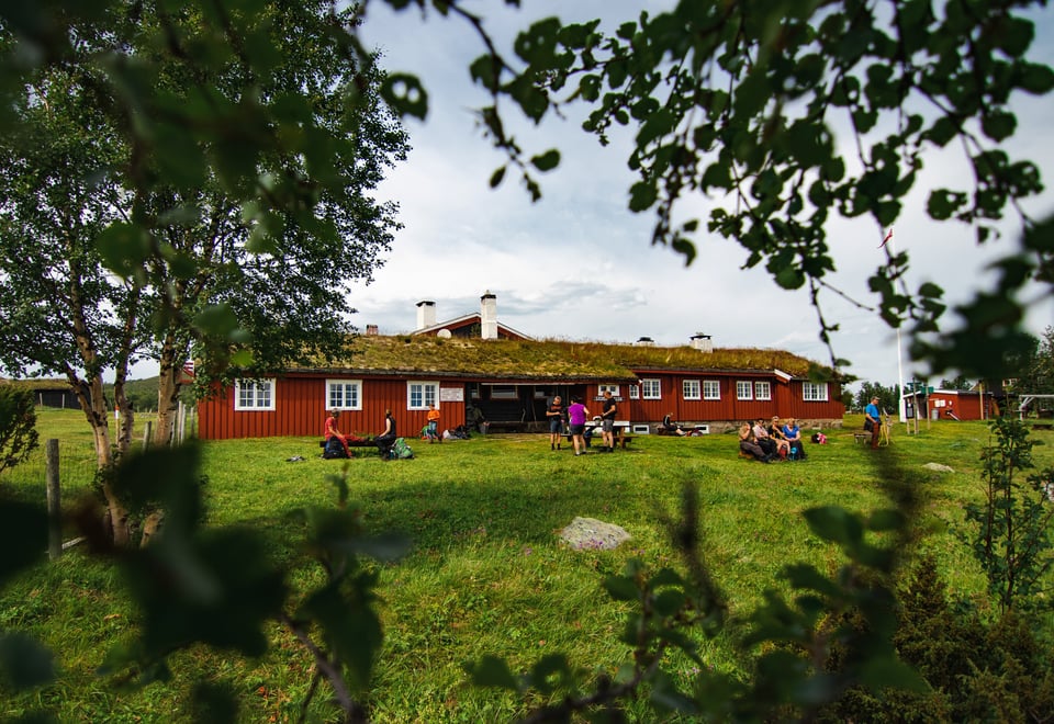 En rød og langstrakt hytte, med et gressdekket landskap foran seg, og noen trær mellom oss og bygningen