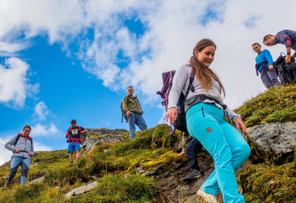 En gjeng med unge voksne på tur i fjellet. Helt foran ser du en ung jente med turkise bukser som smiler og går ned en skråning.