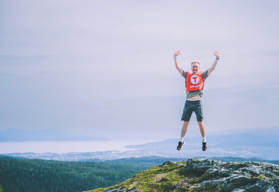 En mann med en rød vest med T for turistforeningen på hopper med armene i været og smiler på en fjelltopp. I bakgrunnen kan man skimte byen Trondheim.