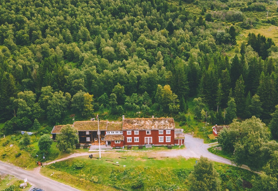 Dronebilde av en turisthytte, sett på skrå nedover ovenfra. Rød bygning med hvite kanter, omgitt av tykk skog på tre sider, og en vei som går forbi foran gressplenen.
