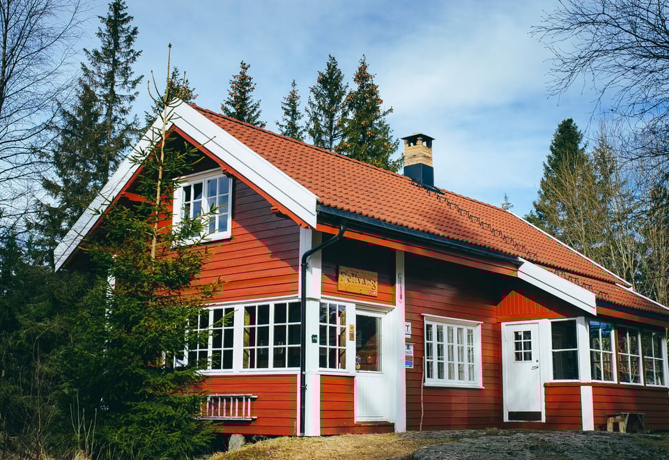 Rødmalt hytte med hvite karmer og oransje takstein i barskog. 