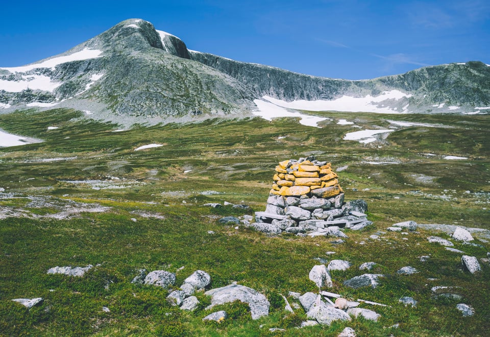 En åpen grønn slette med en varde av stablede steiner, hvor øverste halvdel av steiene har blitt malt gule. I bakgrunnen er et lite fjell.