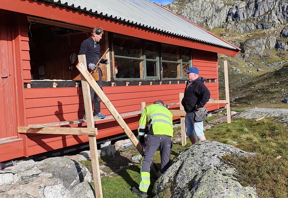 Tre menn står utenfor en hytte og bygger med planker foran en rød hytte. Det mangler et vindu i hytten.
