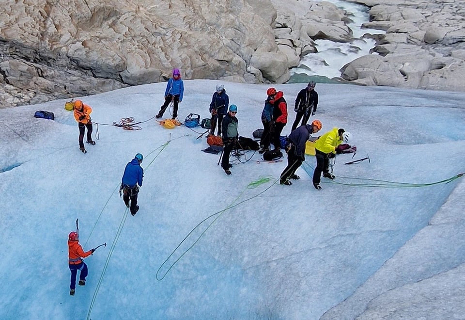 En gruppe mennesker klatrer oppover en blå isbre. Mye grå stein og en liten foss i bakgrunnen.