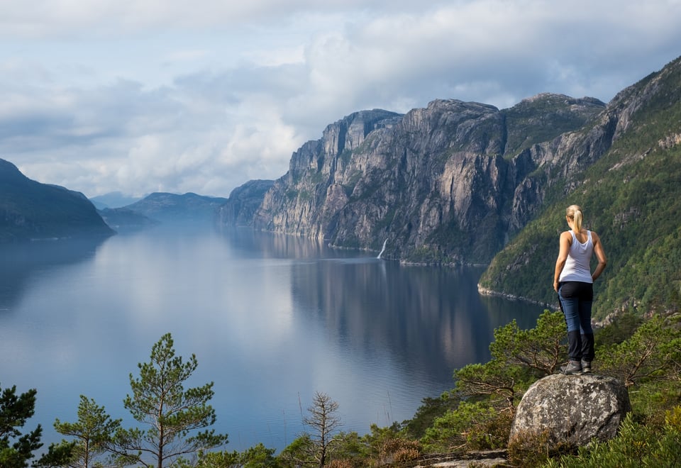 En dame med lys hestehale, iført hvit singlet og blå bukse står på et fjell og ser utover fjorden.