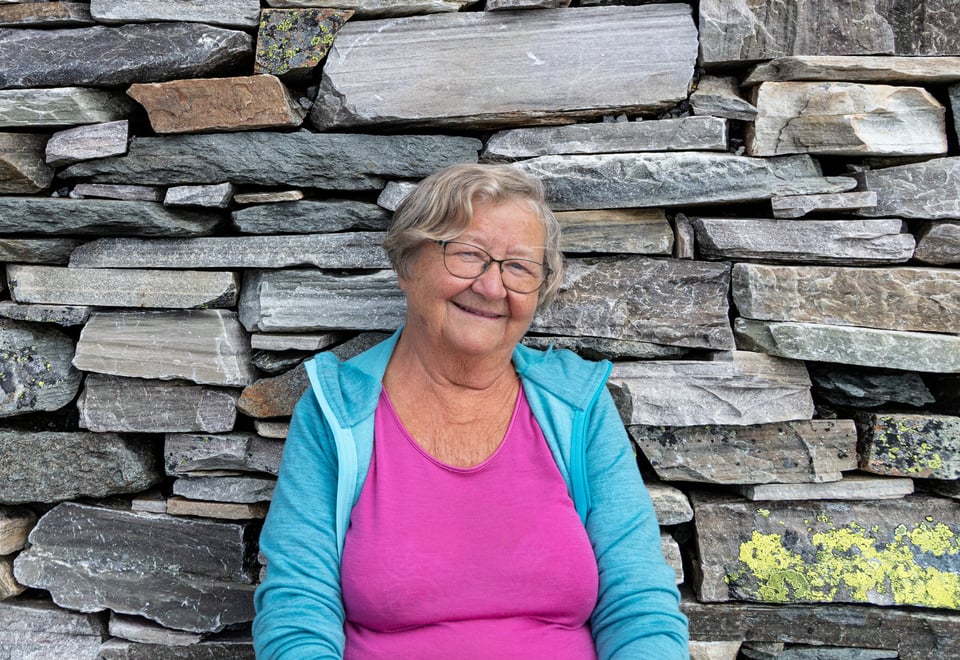 En smilende kvinnelig hyttevert sitter inntil en steinvegg og ser i kamera.
