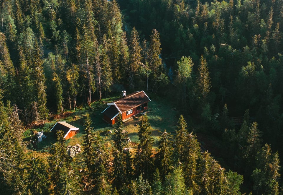 Luftfoto av en liten, rød trehytte med et hvitt skorsteinsrør, omgitt av tett skog i Nordmarka.