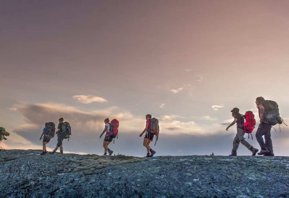 Seks personer med tursekk på er sett fra siden, idet de går langs kanten av en steinrygg. i kvelds-lys. 