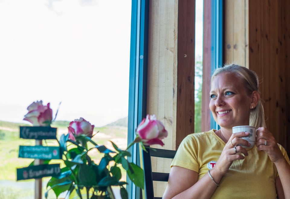 En kvinne som sitter ned i låven og drikker kaffe mens hun speider ut av vinduet. Vase med rosebukett i front.