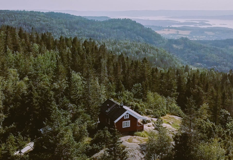 Rød hytte med hvite vinduer omkranset av tett skog og åser.