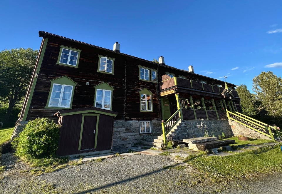 En to-etasjes brun hytte med stor veranda foran, sett fra froskeperspektiv. Skyfritt sommervær.