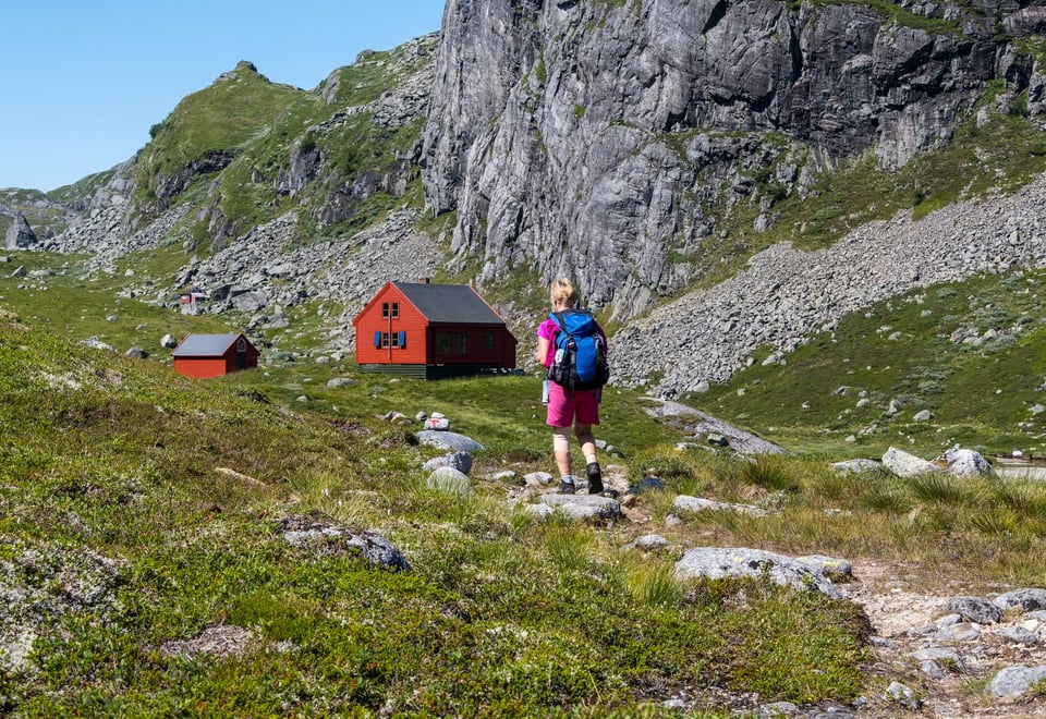 En kvinne i rosa turklær og blå sekk går mot en rød hytte i grønt terreng.