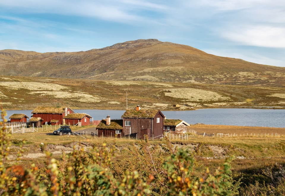 Bildet viser en liten hytte ved siden av en fjellseter i relativt flatt fjellterreng, et fjell og en stor innsjø i bakgrunnen. Fint vær med sol, og antydning til høstfarger på vegetasjon.