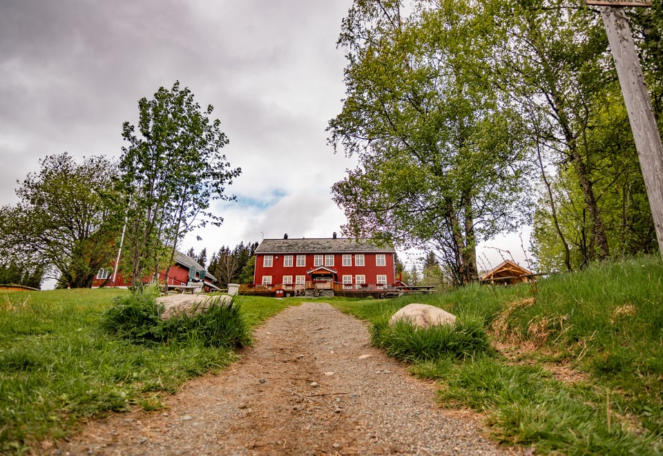 En rød stue med hvite lister og kanter er midt i bildet, og en gjørmete vei leder rett fra synspunktet og opp en bakke mot bygningen. Noen løvtrær står rundt omkring, og bakken er gressdekket. Overskyet vær. 