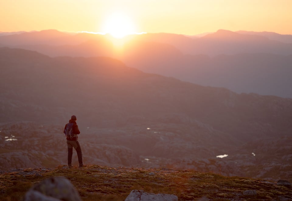 En person speider utover fjelllandskapet med sola som går ned bak fjellene. Oransje lys i bildet.