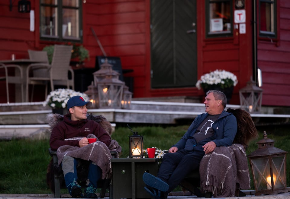 To menn sitter i hver sin stol utendørs i kveldssola og snakker sammen. Lykter med stearinlys er tent.