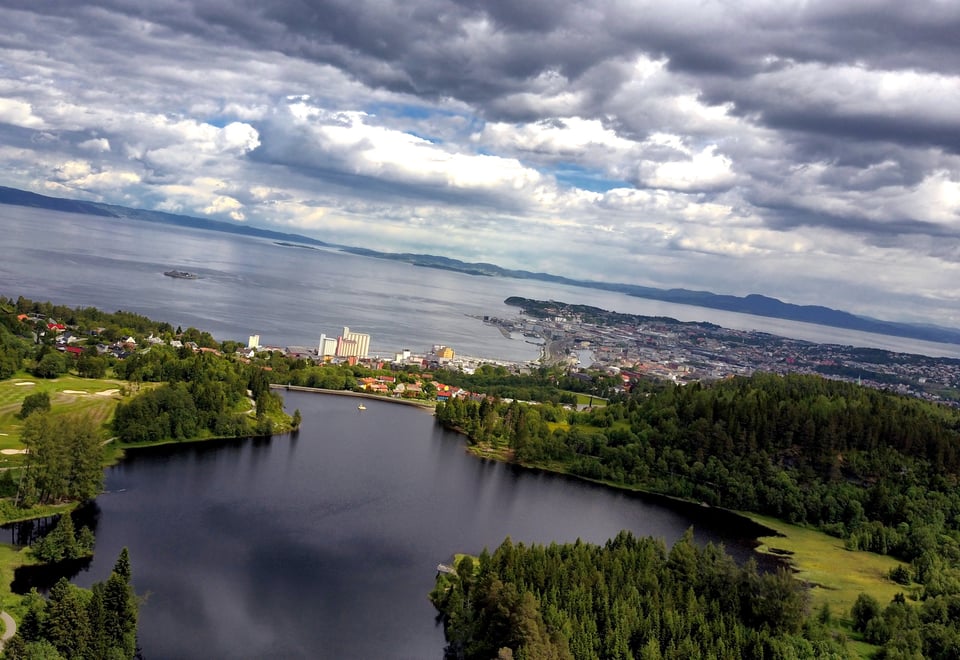 Dronebilde av Theisendammen sett ovenfra. En innsjø i bymarka, med fjorden synlig i bakgrunnen. Skyfull himmel. 