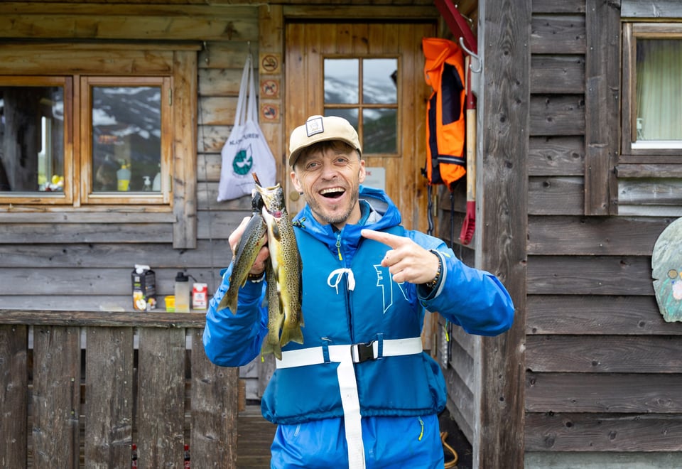 Mann i blå jakke smiler foran hytte og peker på noen fjellørret han har fanget.