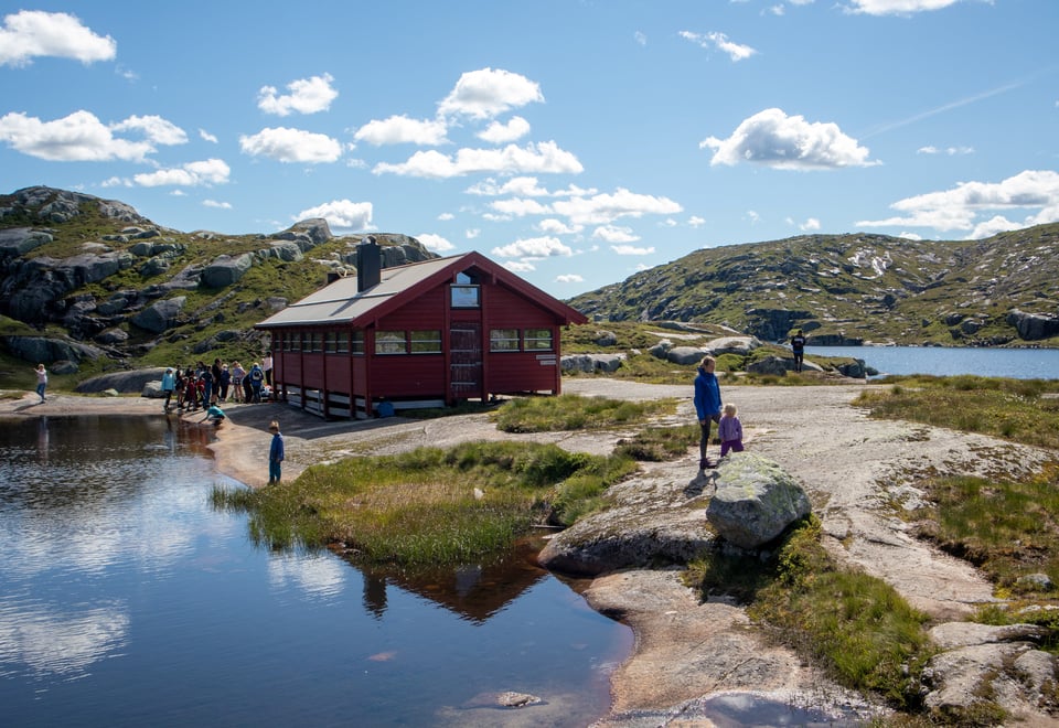 Rød hytte med vann. Barn og voksne utfor hytta. 