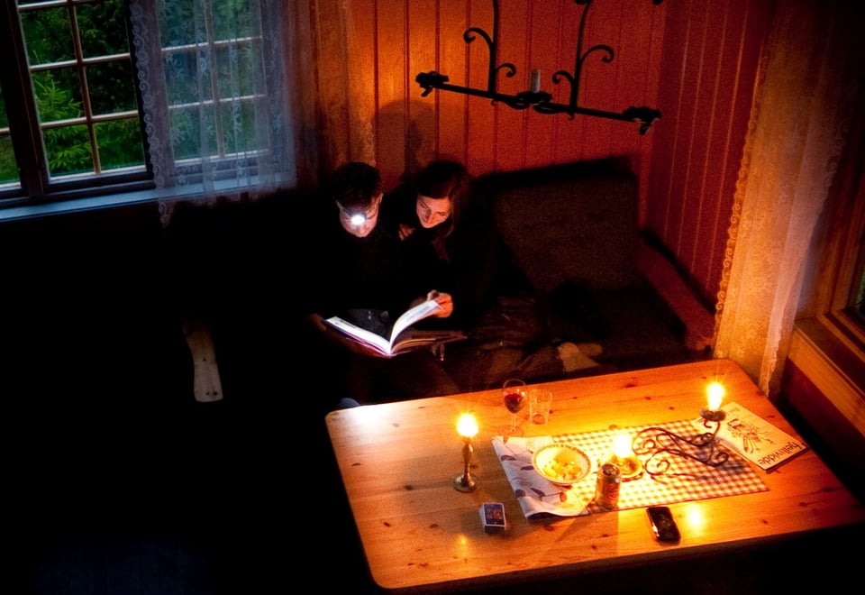 To personer leser en bok sammen i dempet belysning, omgitt av levende lys i en koselig hytte.