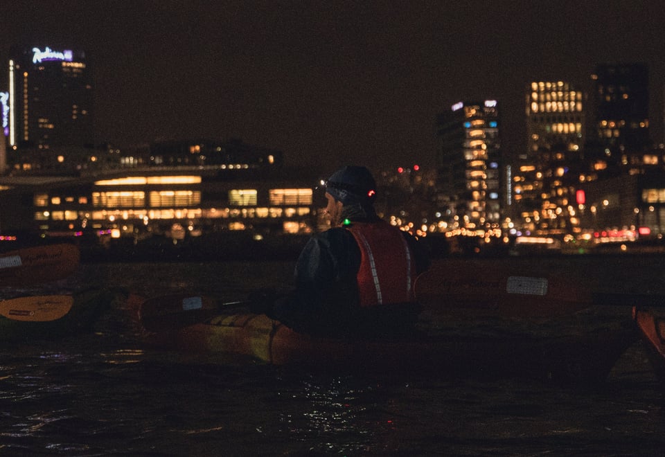 En person i kajakk ses bakfra, iført en rød redningsvest og en hodelykt, mens han padler på vannet om natten. Byens lys reflekteres i vannet, og flere bygninger er synlige i bakgrunnen, inkludert høye tårn og opplyste vinduer, noe som gir en urban og atmosfærisk bakgrunn.