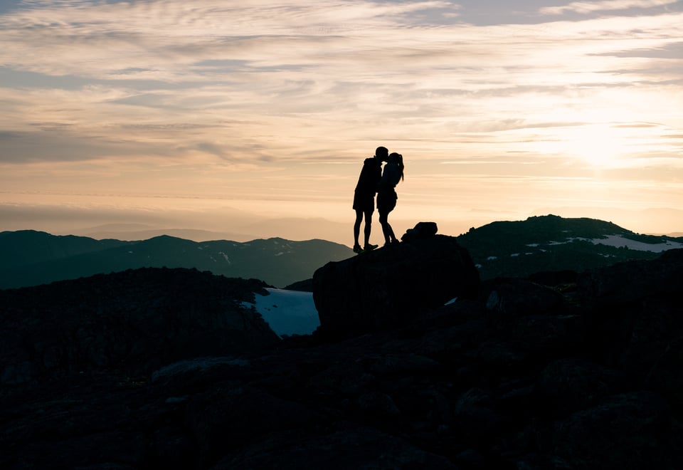 Kjærestepar kysser i solnedgang på fjelltopp.