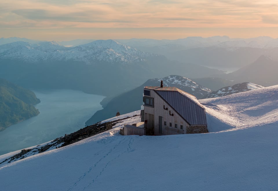 Langstrakt hytte på toppen av fjell i solnedgang med fjord lenger bak.