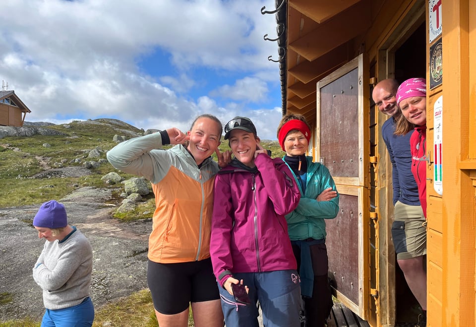 Et turfølge med menn og damer står utenfor en gul hytte og smiler til kamera.