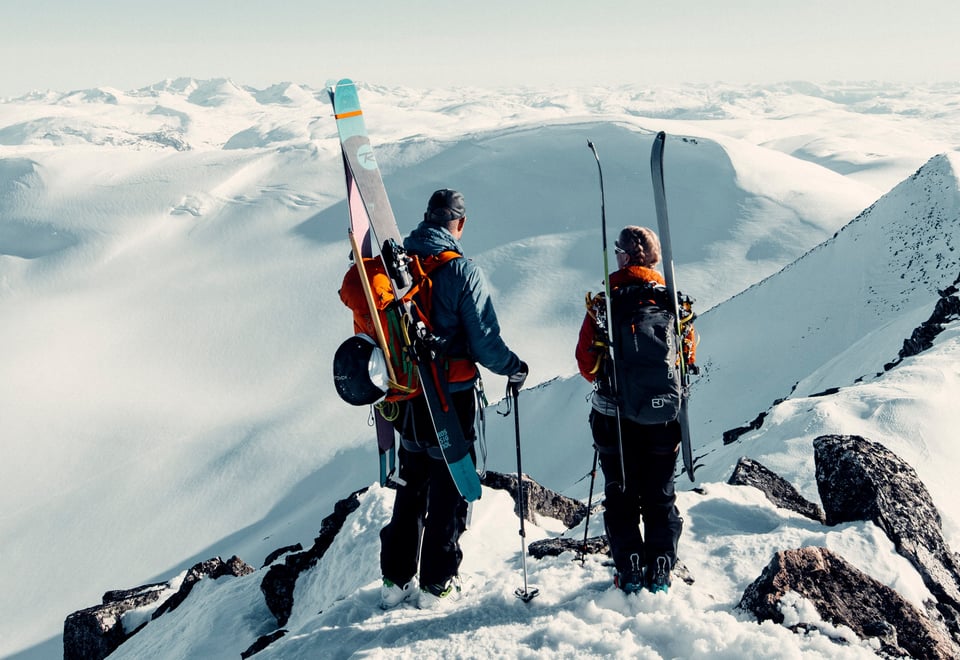 To personer med sekker hvor det er festet på ski på. De står og ser utover et snødekket landskap