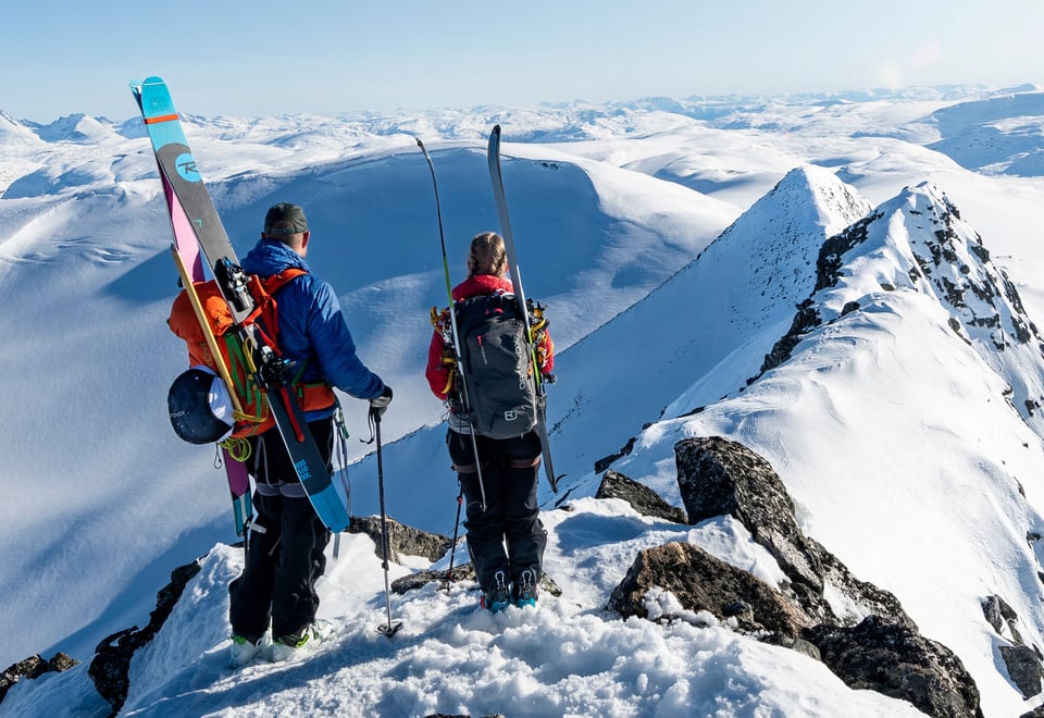 To personer står på en snødekt fjelltopp med skiutstyr, og ser utover et storslått vinterlandskap med fjell i horisonten.