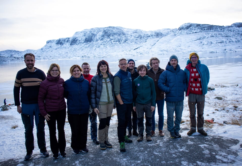 En gruppe med mennesker som står samlet og smiler til kamera. Bak dem er det fjell og vann som er is og snødekket.